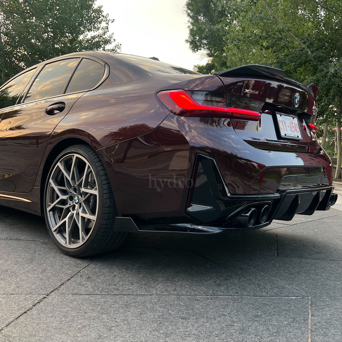 Rear three-quarter view of a dark metallic brown BMW sedan parked outdoors on a paved surface with trees in the background. The image highlights a black rear diffuser and lip spoiler kit installed on the vehicle.