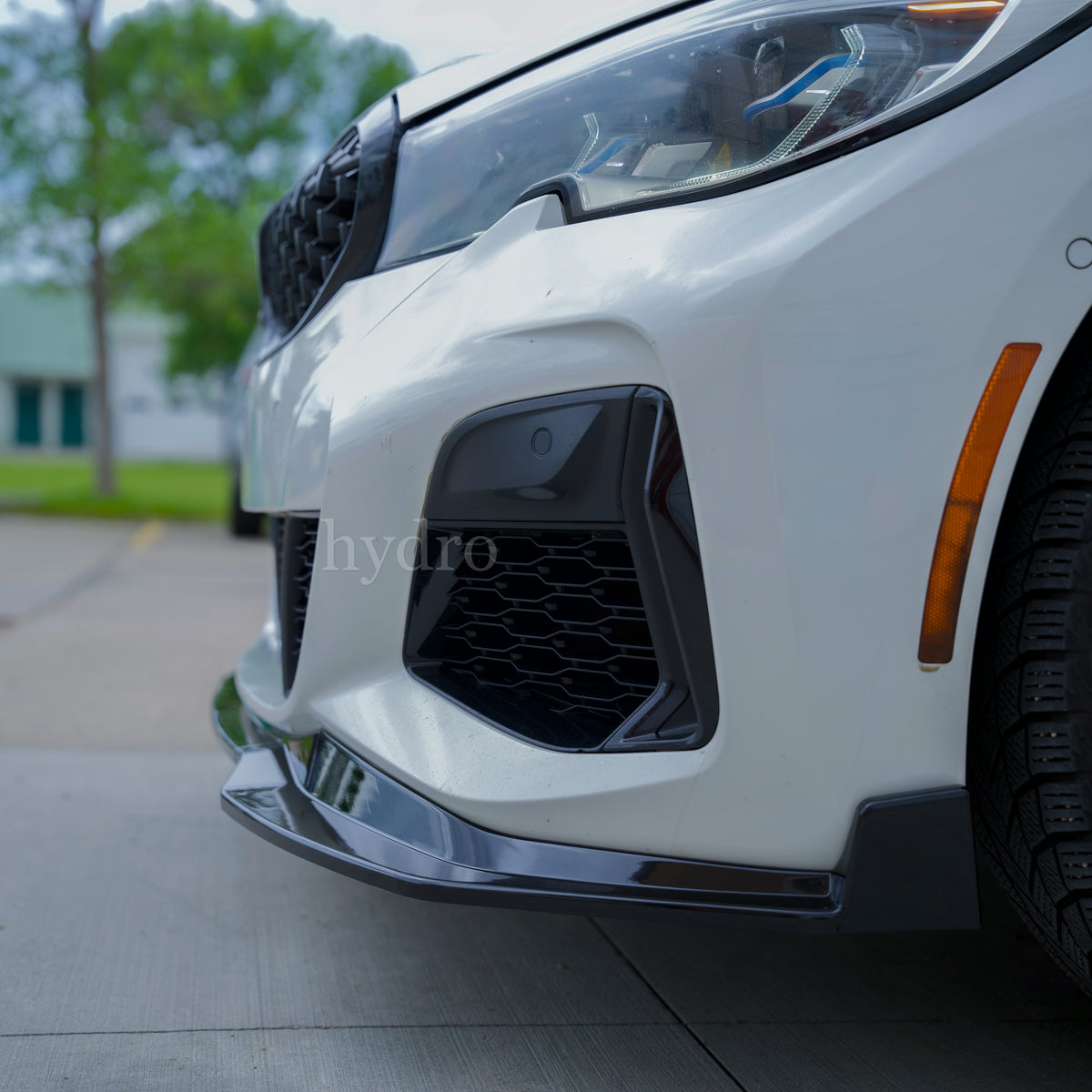Front bumper and lower front splitter of a white car, showing black honeycomb grille inserts and a glossy black lip spoiler on a paved outdoor surface.